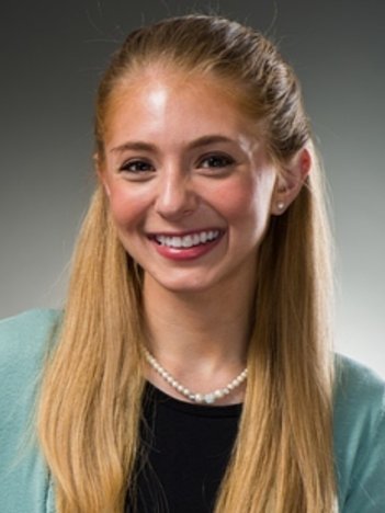 A white woman with blonde hair wears a black blouse, cyan cardigan, and pearl necklace and smiles in front of a gray background.